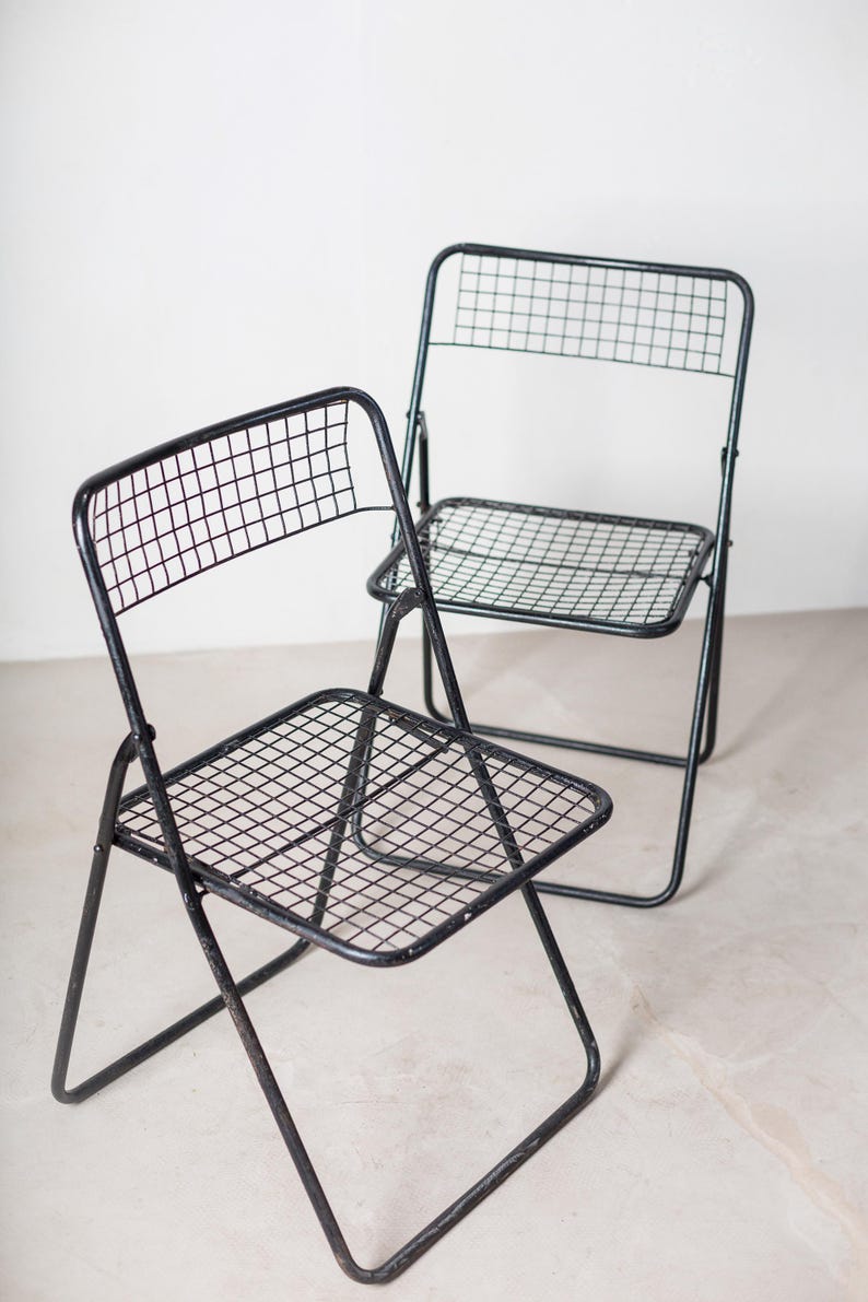 May include: Two black metal folding chairs with a wire mesh seat and back. The chairs are in a white room.