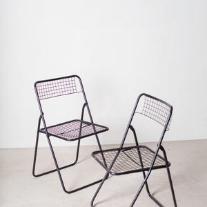 May include: Two black metal folding chairs with a wire mesh seat and back. The chairs are folded and sitting on a white floor.