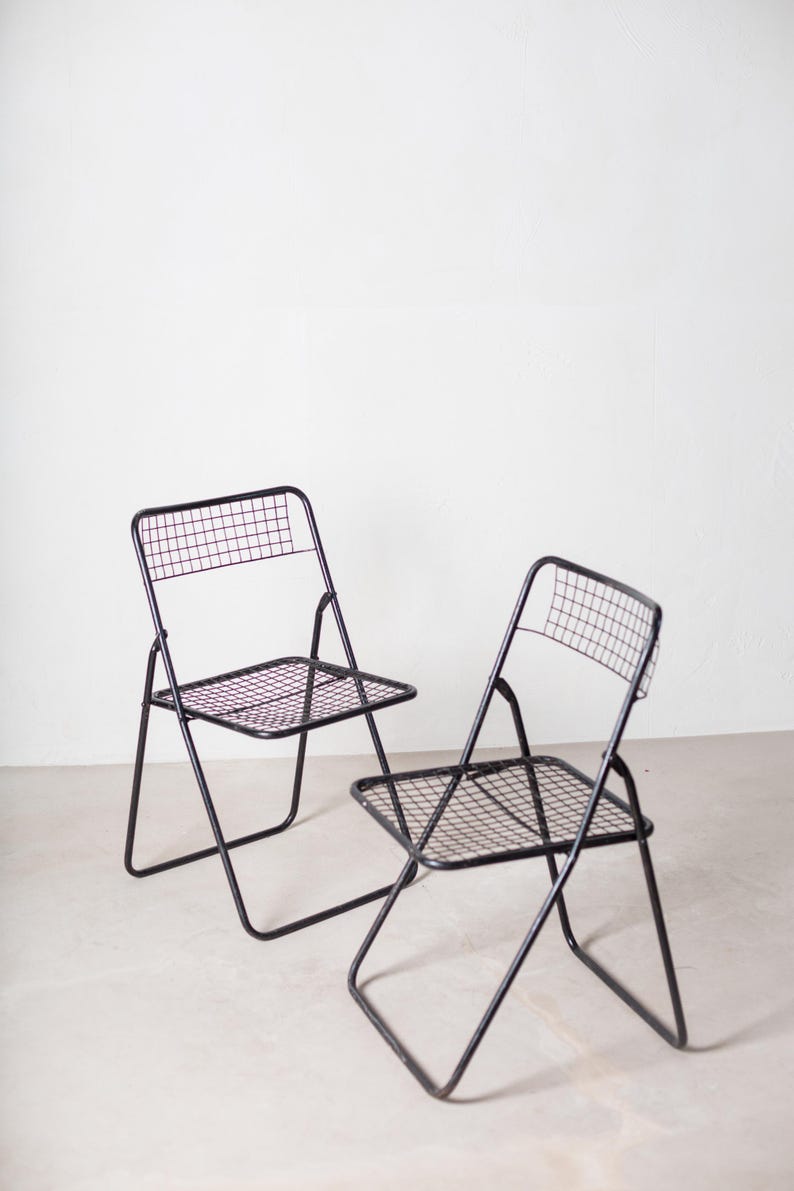 May include: Two black metal folding chairs with a wire mesh seat and back. The chairs are folded and sitting on a white floor.