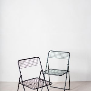 May include: Two black metal folding chairs with a wire mesh seat and back. The chairs are in a white room with a wooden beam in the background.