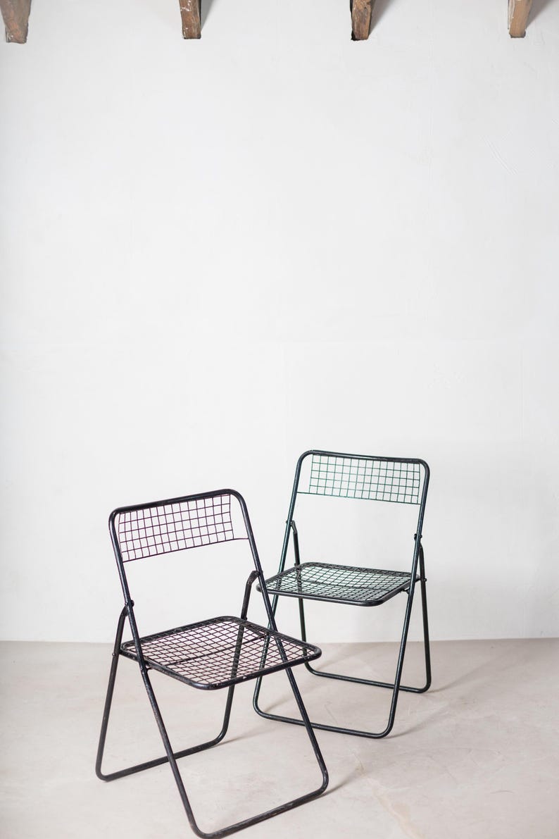 May include: Two black metal folding chairs with a wire mesh seat and back. The chairs are in a white room with a wooden beam in the background.
