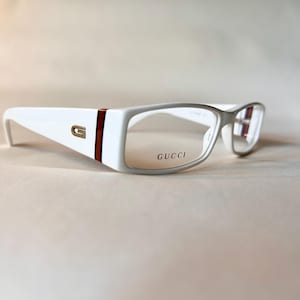 May include: White rectangular eyeglasses featuring a gold-tone G logo on the temple, accented by a vertical red and brown stripe. The right lens is etched with the word GUCCI in black lettering, and the frame has a smooth, glossy finish.