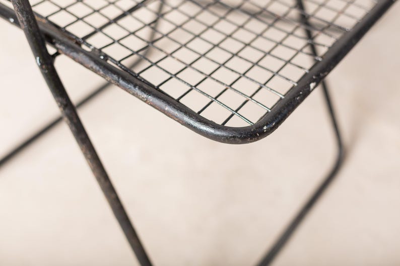 May include: Close-up of a black metal chair with a wire mesh seat. The chair is made of sturdy metal and has a simple, industrial design.