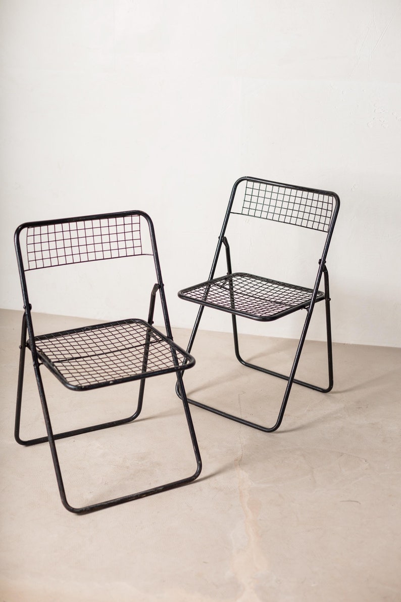 May include: Two black metal folding chairs with a wire mesh seat and back. The chairs are in a light-colored room.