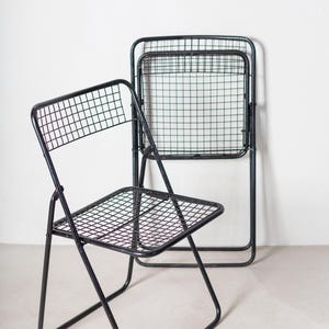 May include: Two black metal folding chairs with a wire mesh seat and back. The chairs are folded and stacked on top of each other.