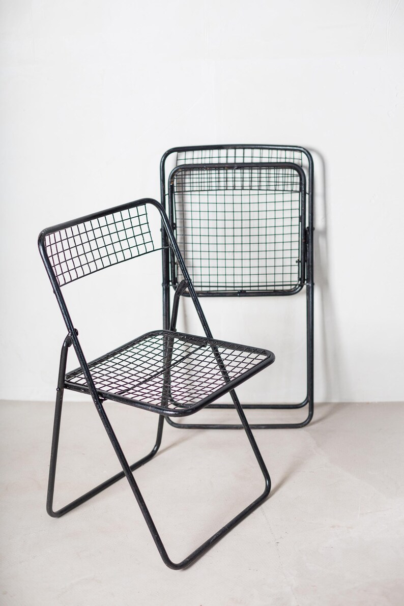 May include: Two black metal folding chairs with a wire mesh seat and back. The chairs are folded and stacked on top of each other.