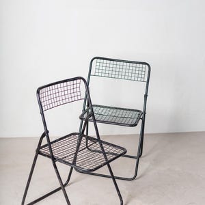 May include: Two black metal folding chairs with a wire mesh seat and back. The chairs are folded and sitting on a light gray floor.