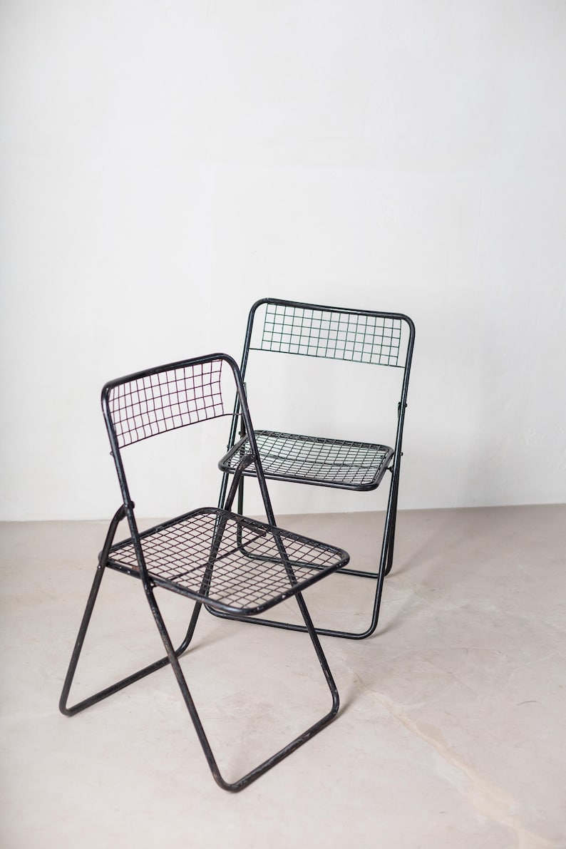 May include: Two black metal folding chairs with a wire mesh seat and back. The chairs are folded and sitting on a light gray floor.