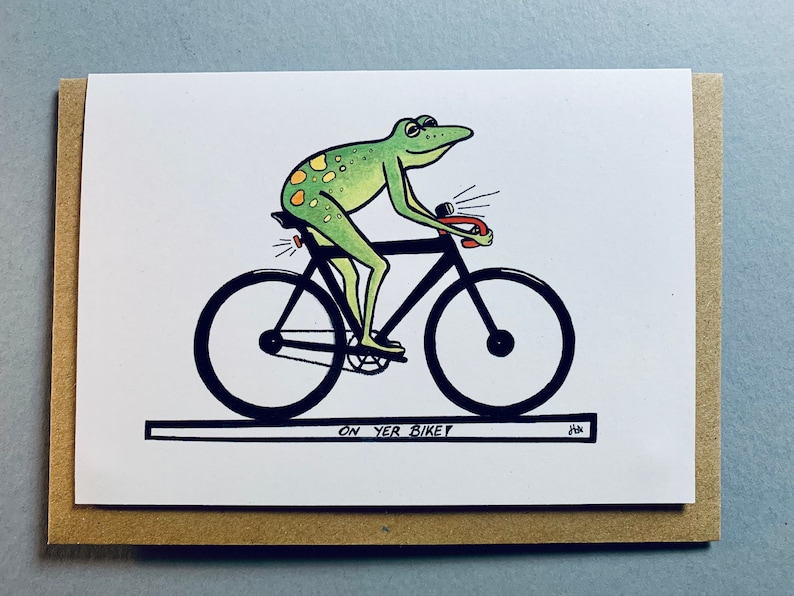 May include: A green frog with yellow spots riding a black bicycle with a red light. The frog is wearing a black helmet and has a black handlebar. The bicycle has a black seat and black wheels. The text "ON YER BIKE" is written below the bicycle.