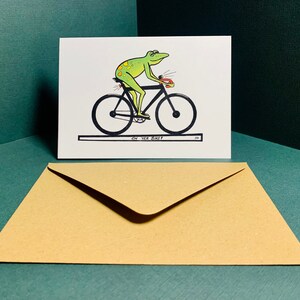 May include: A greeting card with a green frog riding a bicycle. The card has the text "On your bike!" printed on it.