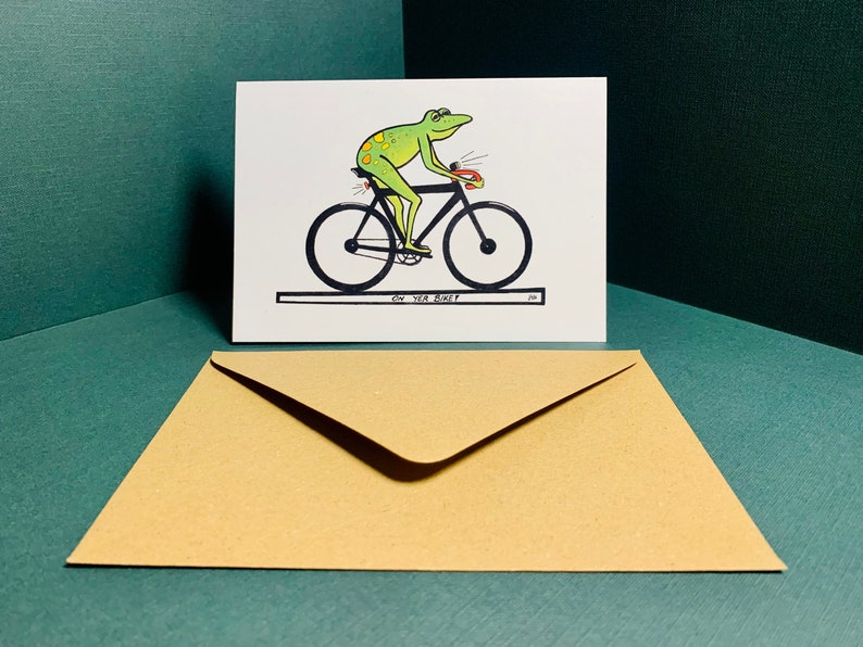 May include: A greeting card with a green frog riding a bicycle. The card has the text "On your bike!" printed on it.