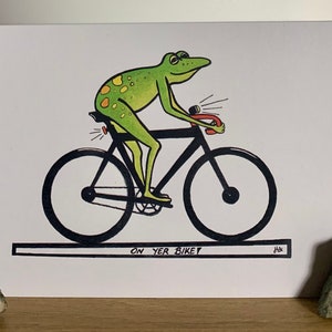 May include: A green frog with yellow spots rides a black bicycle with a red handlebar. The frog has a light on the back of the bike and another light on the front. The text "ON YER BIKE" is written below the bike.