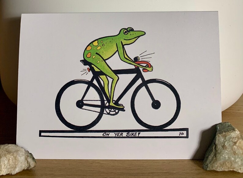 May include: A green frog with yellow spots rides a black bicycle with a red handlebar. The frog has a light on the back of the bike and another light on the front. The text "ON YER BIKE" is written below the bike.