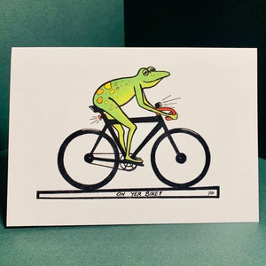 May include: A green frog with yellow spots rides a black bicycle with a red light on the back and a red light on the front. The frog is wearing a black helmet. The bicycle has black wheels and a black seat. The text "ON YER BIKEY" is written below the bicycle.