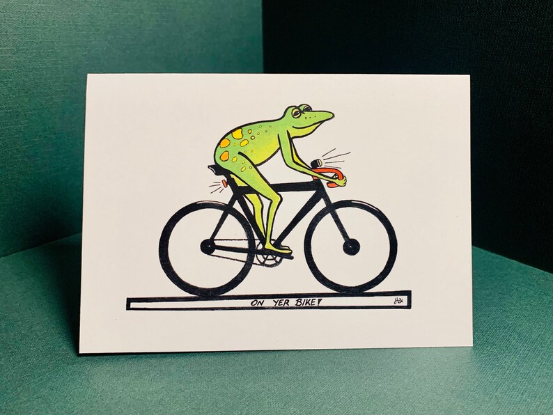 May include: A green frog with yellow spots rides a black bicycle with a red light on the back and a red light on the front. The frog is wearing a black helmet. The bicycle has black wheels and a black seat. The text "ON YER BIKEY" is written below the bicycle.