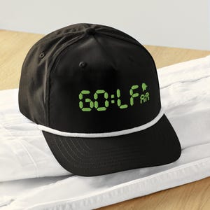Puede incluir: Gorra de golf negra con la palabra "GOLF" en fuente digital verde. La gorra tiene un detalle de cuerda blanca a lo largo del borde y el texto "60:LF am" en verde. La gorra está sobre una superficie blanca.