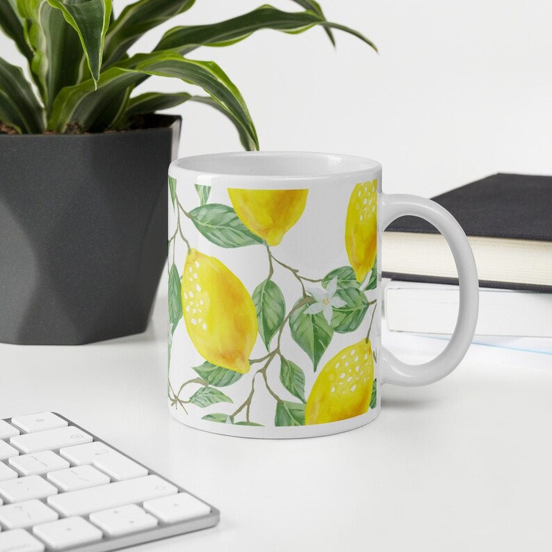 May include: White ceramic mug with a lemon and leaf pattern. The mug has a handle and is filled with a white liquid.