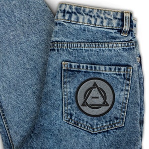 May include: A pair of blue denim jeans with a black and grey patch on the back pocket. The patch features a circle with a triangle inside, and the letter 'A' is inside the triangle.