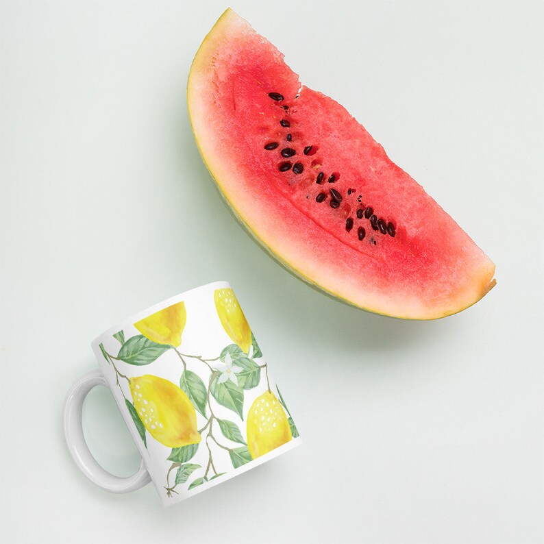 May include: White ceramic mug with a lemon print design. The mug has a white handle and is sitting on a white surface. A slice of watermelon is in the background.