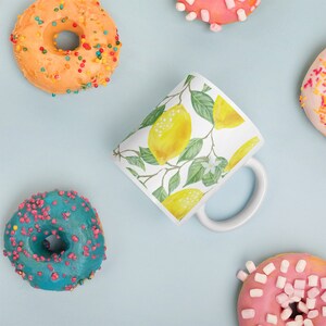 May include: A white ceramic mug with a lemon print design. The mug has a handle and is surrounded by colorful donuts.