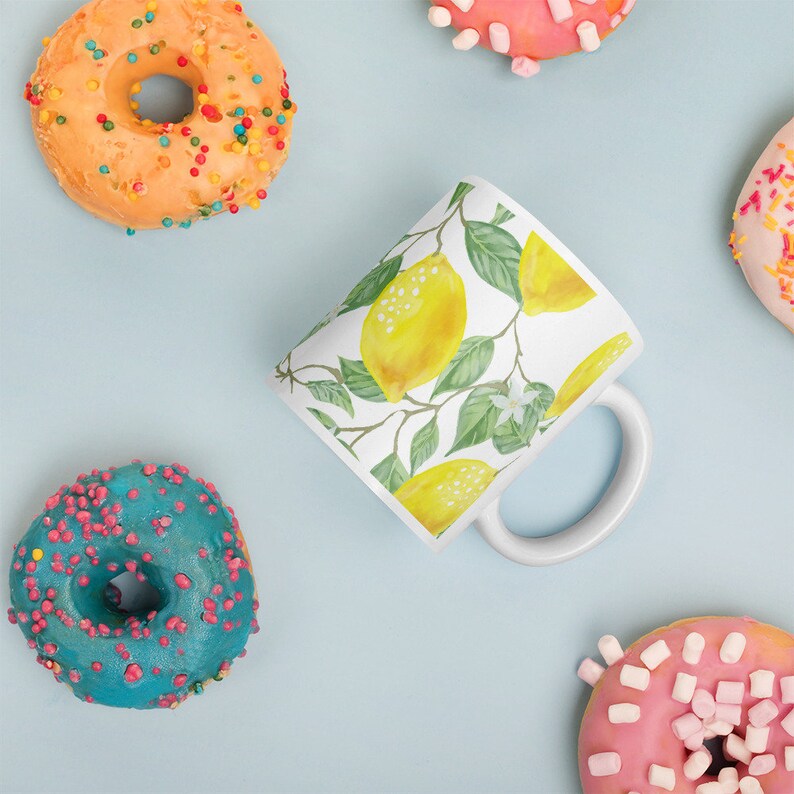 May include: A white ceramic mug with a lemon print design. The mug has a handle and is surrounded by colorful donuts.