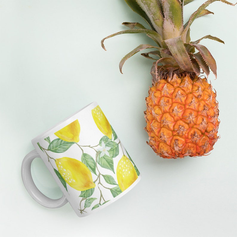 May include: White ceramic mug with a lemon and leaf design. The mug has a white handle. A pineapple is in the background.