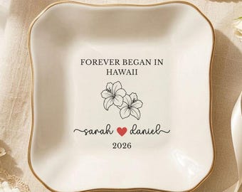 May include: A square white ceramic trinket dish with gold-toned edges. The center features black text reading FOREVER BEGAN IN HAWAII, a line drawing of two hibiscus flowers, the names sarah and daniel with a red heart, and the year 2026.
