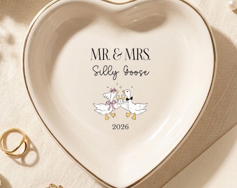 May include: A heart-shaped ceramic trinket dish with a gold-toned rim. The center features the text "MR. & MRS. Silly Goose" above an illustration of two geese clinking glasses, with the year "2026" printed at the bottom in black ink.