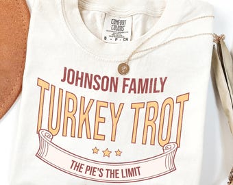 May include: Cream-colored t-shirt with the text "JOHNSON FAMILY TURKEY TROT" in orange and maroon. The phrase "THE PIE'S THE LIMIT" is below the main text, with "EST. 2025" underneath. A gold necklace and a brown hat are also in the image.