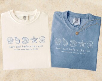 May include: Two folded t-shirts, one white and one blue, with a seashell design and the text "last sail before the veil santa rosa beach, 2026". A gold necklace rests on the blue shirt.