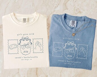 May include: Two t-shirts, one cream and one blue, are displayed. Each shirt features the text "girls gone mild" and a graphic of a sleep mask, heart-shaped glasses, and champagne flutes. The text "sarah's bachelorette san diego, ca 2026" is also printed on the shirts.