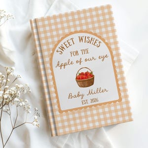 Könnte beinhalten: Ein gebundenes Buch mit einem hellorange-weißen Vichy-Karo-Muster auf dem Einband. In der Mitte befindet sich ein weißes Etikett mit Wellenrand und dem Text "SWEET WISHES FOR THE Apple of our eye", einem Korb mit roten Äpfeln, "Baby Miller" und "EST. 2026".