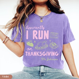 K&ouml;nnte beinhalten: Ein violettes Kurzarm-T-Shirt mit dem Text "Apparently I RUN on THANKSGIVING" in Pink und Wei&szlig;. Die Grafik zeigt ein St&uuml;ck Kuchen, einen gebratenen Truthahn, ein Paar Laufschuhe und eine Marathon-Startnummer 15432 in hellem Gr&uuml;n.
