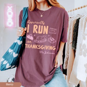 K&ouml;nnte beinhalten: Ein beerenfarbenes T-Shirt mit dem Text "Apparently I RUN on THANKSGIVING" in hellem Orange sowie grafischen Illustrationen eines St&uuml;cks Kuchen, eines gebratenen Truthahns, eines Laufschuhpaars und einer Marathon-Startnummer "15432".