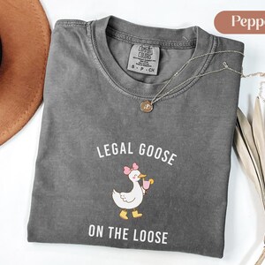 Puede incluir: Una camiseta gris Comfort Colors con la frase "LEGAL GOOSE ON THE LOOSE" y un dibujo de un ganso con un lazo rosa y una bebida. Un collar dorado est&aacute; sobre la camiseta.
