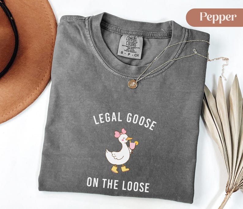Puede incluir: Una camiseta gris Comfort Colors con la frase "LEGAL GOOSE ON THE LOOSE" y un dibujo de un ganso con un lazo rosa y una bebida. Un collar dorado est&aacute; sobre la camiseta.