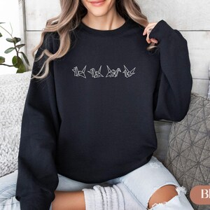 May include: Black crewneck sweatshirt with a white origami crane design embroidered across the chest. The design features five stylized cranes in a row. The sweatshirt is made of a soft material and is suitable for casual wear.
