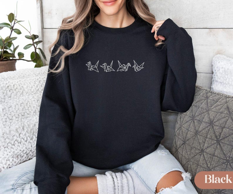 May include: Black crewneck sweatshirt with a white origami crane design embroidered across the chest. The design features five stylized cranes in a row. The sweatshirt is made of a soft material and is suitable for casual wear.