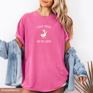 Puede incluir: Camiseta rosa brillante con el texto "LEGAL GOOSE ON THE LOOSE" y un dibujo de un ganso. La camiseta es de color liso y tiene mangas cortas. Es de estilo casual.