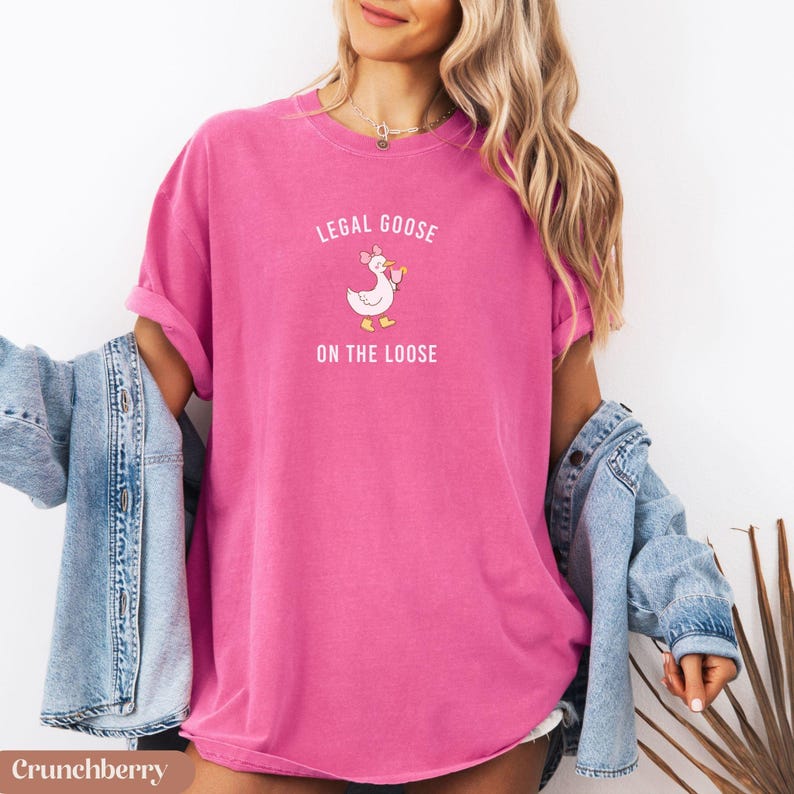 Puede incluir: Camiseta rosa brillante con el texto "LEGAL GOOSE ON THE LOOSE" y un dibujo de un ganso. La camiseta es de color liso y tiene mangas cortas. Es de estilo casual.