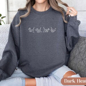 May include: Dark heather gray sweatshirt with a white origami crane design. The long-sleeved crew neck top is made of a soft material. The text "Dark Heather" is at the bottom of the image.