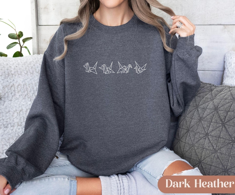 May include: Dark heather gray sweatshirt with a white origami crane design. The long-sleeved crew neck top is made of a soft material. The text "Dark Heather" is at the bottom of the image.