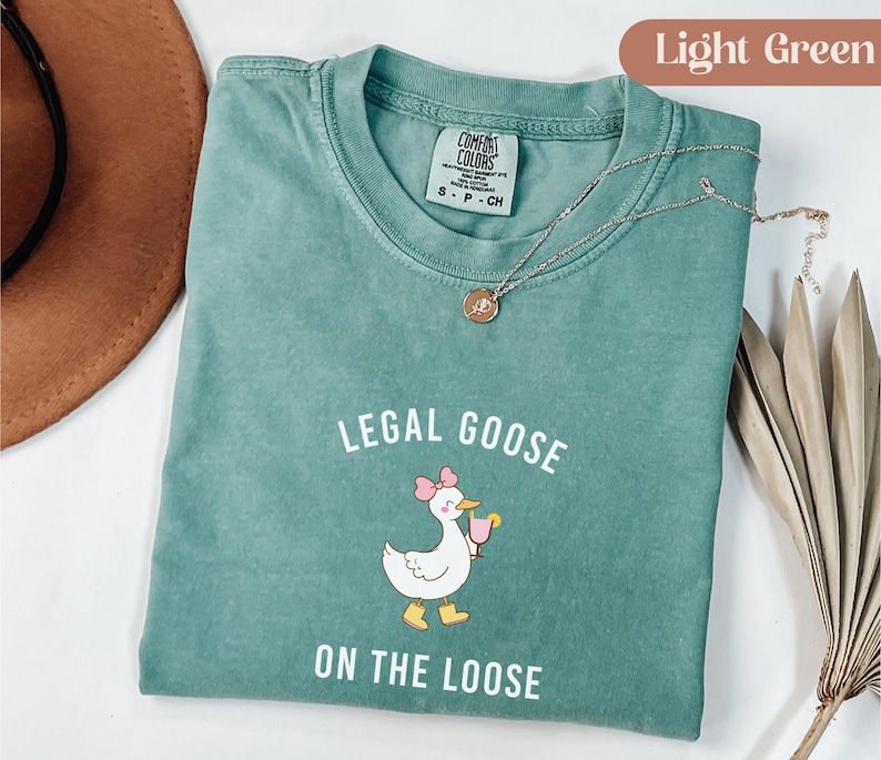 Puede incluir: Camiseta verde claro con la frase "LEGAL GOOSE ON THE LOOSE" y un dibujo de un ganso con una bebida. La camiseta tiene cuello redondo y mangas cortas. Un collar plateado con un colgante est&aacute; sobre la camiseta.