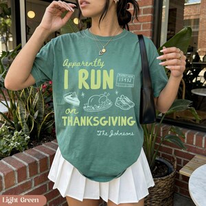 K&ouml;nnte beinhalten: Ein kurz&auml;rmeliges T-Shirt in Salbeigr&uuml;n mit dem hellgr&uuml;nen Schriftzug "Apparently I RUN on THANKSGIVING". Die Grafik zeigt ein St&uuml;ck Kuchen, einen gebratenen Truthahn, ein Paar Turnschuhe, eine Startnummer "15432" und "The Johnsons".