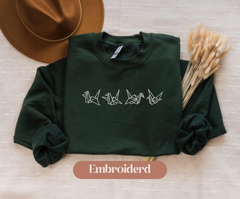 May include: A dark green sweatshirt with white embroidered origami cranes. The sweatshirt has long sleeves and a crew neck. The word "Embroidered" is printed on a brown oval at the bottom of the image.
