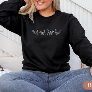 May include: Black crew neck sweatshirt with a white origami crane design. The design features four stylized cranes in a row. The sweatshirt is a solid black color and has long sleeves. The model is wearing blue jeans.