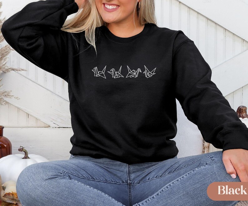 May include: Black crew neck sweatshirt with a white origami crane design. The design features four stylized cranes in a row. The sweatshirt is a solid black color and has long sleeves. The model is wearing blue jeans.