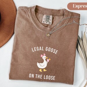 Puede incluir: Camiseta color espresso con el texto "LEGAL GOOSE ON THE LOOSE" y un dibujo de un ganso con un lazo rosa y una bebida. La camiseta tiene cuello redondo y una peque&ntilde;a etiqueta. Un collar plateado est&aacute; sobre la camiseta.