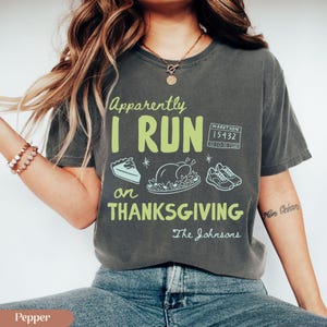K&ouml;nnte beinhalten: Ein anthrazitfarbenes Kurzarm-T-Shirt mit dem hellgr&uuml;nen Text Apparently I RUN on THANKSGIVING und wei&szlig;en Strichzeichnungen eines Tortenst&uuml;cks, eines gebratenen Truthahns, eines Laufschuhpaars sowie einer Startnummer mit der Aufschrift 15432.