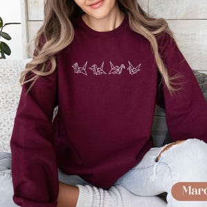 May include: Maroon-colored sweatshirt featuring a minimalist design of four origami cranes in white thread. The sweatshirt has a crew neck and long sleeves. The word "Maroon" is displayed in a small rectangular label at the bottom right.